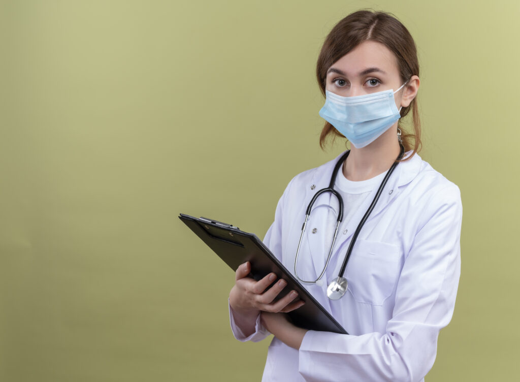 seriously looking young female doctor wearing medical robe mask stethoscope holding clipboard isolated green space with copy space